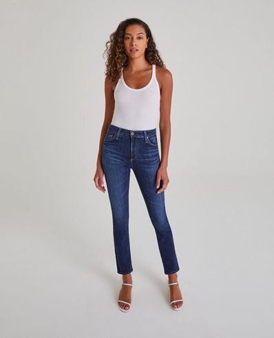 High-waisted slim-fit dark blue jeans worn by a woman with curly hair, stylishly paired with a white tank top and nude heels, showcasing contemporary denim fashion.