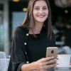 Katyamaker, woman smiling at café while using smartphone, casual fashion, modern lifestyle, customer engagement, bright indoor setting, professional woman in relaxed environment, outdoor cafe scene with coffee cup and table, female entrepreneur, confident and approachable woman.