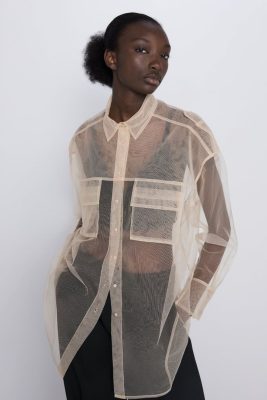 Sheer beige button-up shirt with oversized pockets, lightweight and transparent, styled with black pants, modern and minimalist fashion for women.