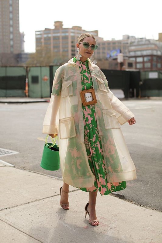 Flowy green and pink patterned dress paired with beige oversized jacket, stylish accessories including sunglasses and earrings, holding green handbag, urban street fashion, ready for spring or summer, modern chic city look.