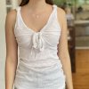 Delicate white linen sleeveless top with tie front detail and knotted shoulder straps, paired with elegant jewelry, set in a cozy home kitchen environment.