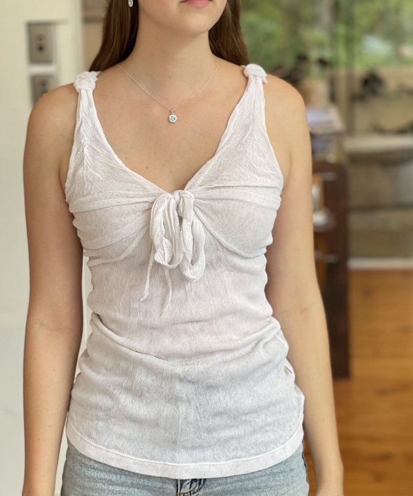 Delicate white linen sleeveless top with tie front detail and knotted shoulder straps, paired with elegant jewelry, set in a cozy home kitchen environment.