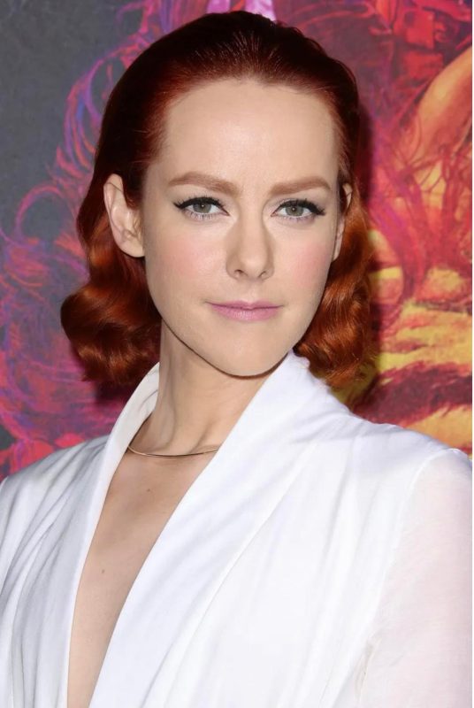 Elegant red-haired woman with wavy hairstyle, wearing white blazer, posing confidently in front of a colorful abstract background, showcasing fashionable portrait photography.