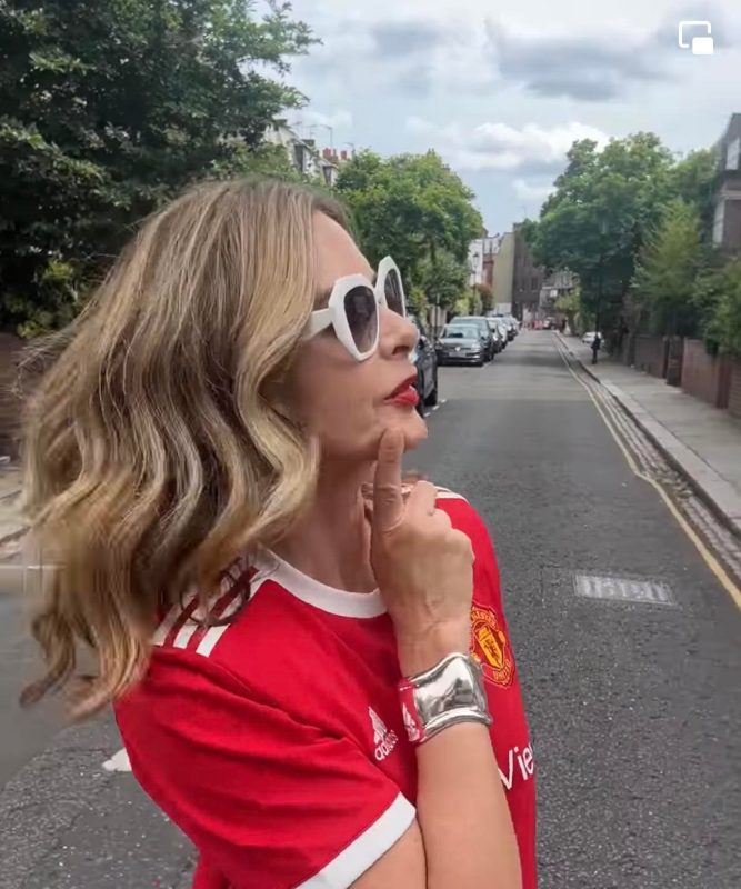 Stylish woman with blonde wavy hair wearing white sunglasses and a red Manchester United jersey, standing on a city street with trees and parked cars, embracing fashion and confident attitude.
