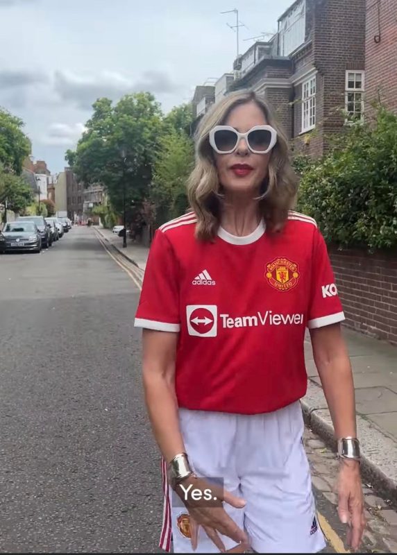 Bold woman wearing vintage white sunglasses and a Manchester United football jersey standing on a city street.