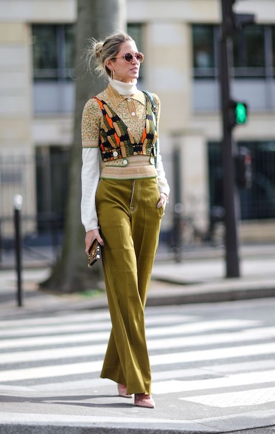 Stylish woman in fashion-forward outfit crossing street wearing trendy wide-leg pants, patterned vest, turtleneck, sunglasses, and carrying a clutch on an urban city background.