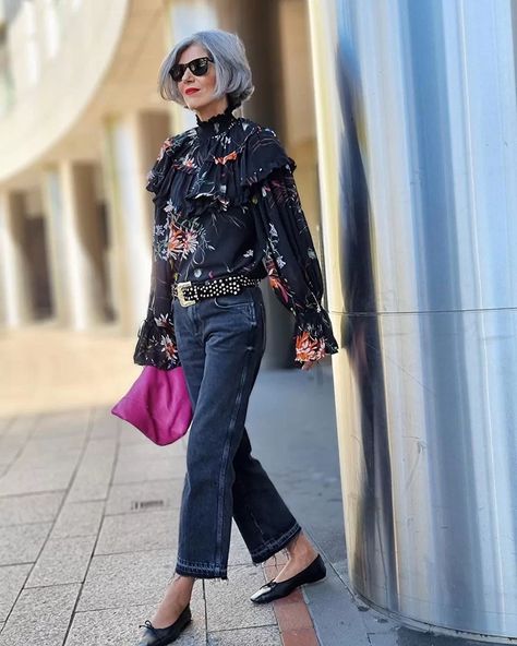 Elegant woman in fashionable floral blouse, dark jeans, and sunglasses standing outdoors with modern architecture background, showcasing trendy street style and personal fashion.