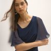 Glamorous woman in navy evening dress with sheer sleeve details and sparkling earrings, posing against white wall, fashion photography for formal wear and jewelry.
