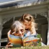Reading a children's adventure book titled "Tina's Great Escape" outdoors on the lawn with a grand Victorian house in the background.