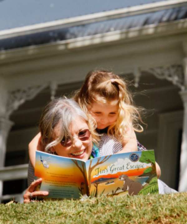Reading a children's adventure book titled "Tina's Great Escape" outdoors on the lawn with a grand Victorian house in the background.