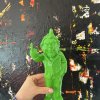 Colorful green resin sculpture of a quirky man with a raised middle finger, set against an abstract textured black background with splashes of red, yellow, and white paint.