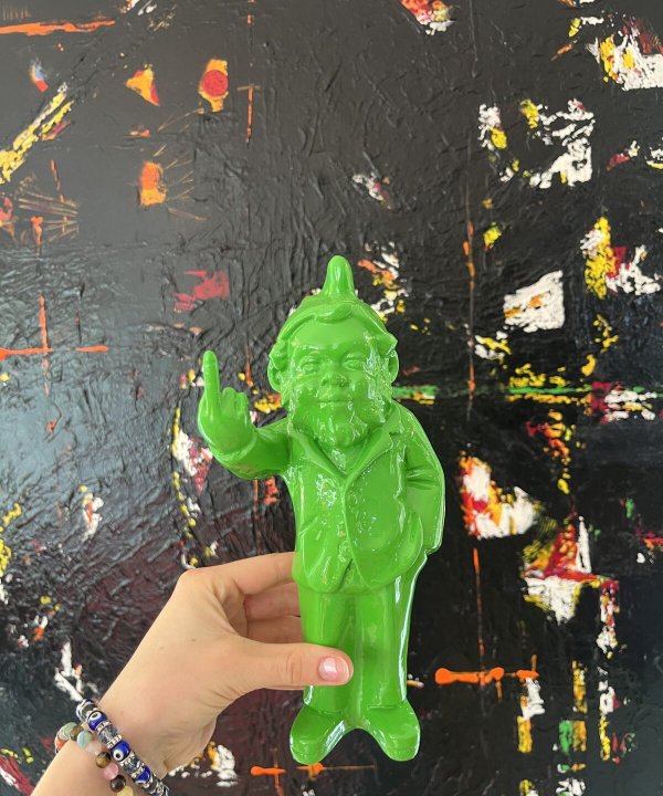 Colorful green resin sculpture of a quirky man with a raised middle finger, set against an abstract textured black background with splashes of red, yellow, and white paint.